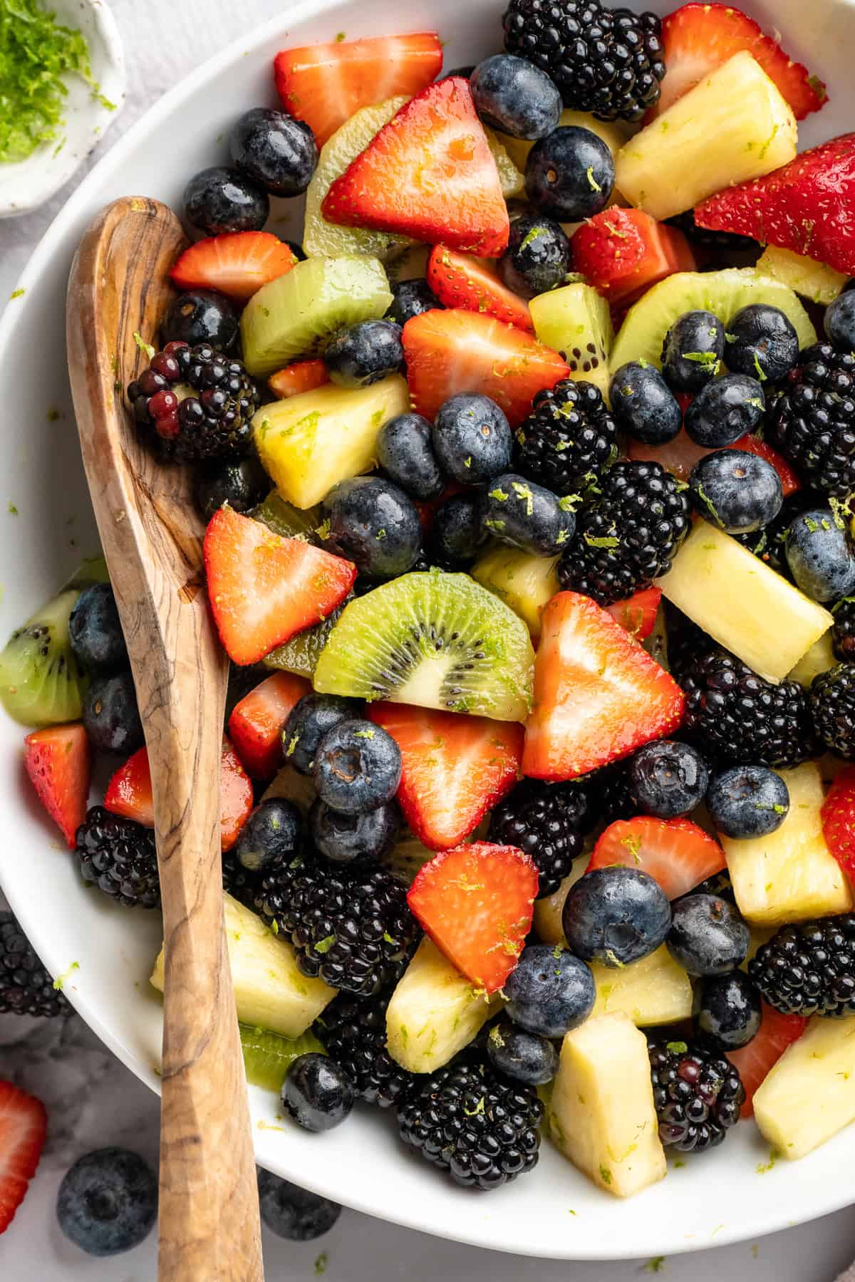This bright rainbow fruit salad is tossed in a fresh maple lime dressing for a side that's juicy, refreshing, and perfect for any occasion! Wooden spoon resting in serving bowl of rainbow fruit salad.