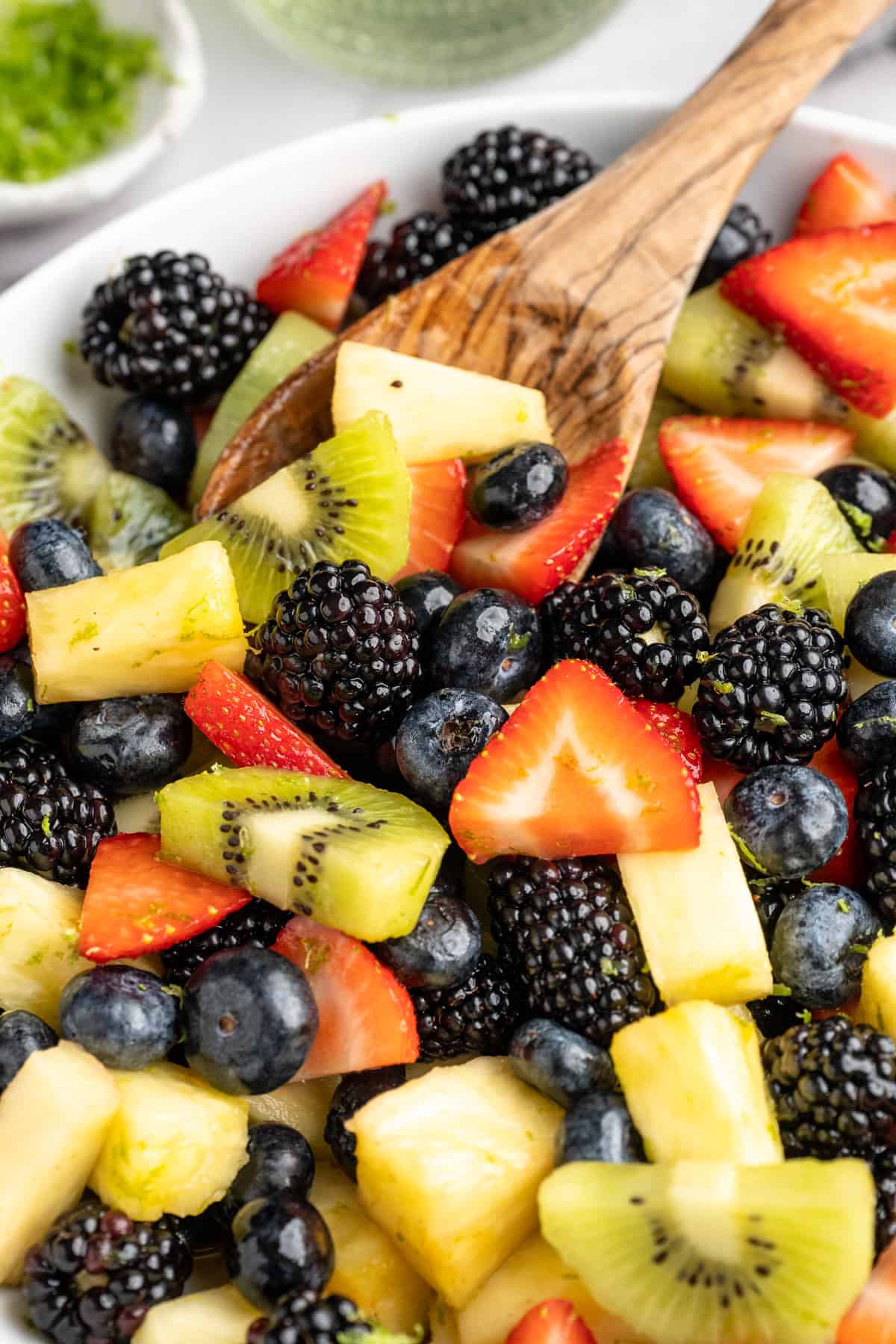 This bright rainbow fruit salad is tossed in a fresh maple lime dressing for a side that's juicy, refreshing, and perfect for any occasion! Wooden spoon in bowl of rainbow fruit salad.