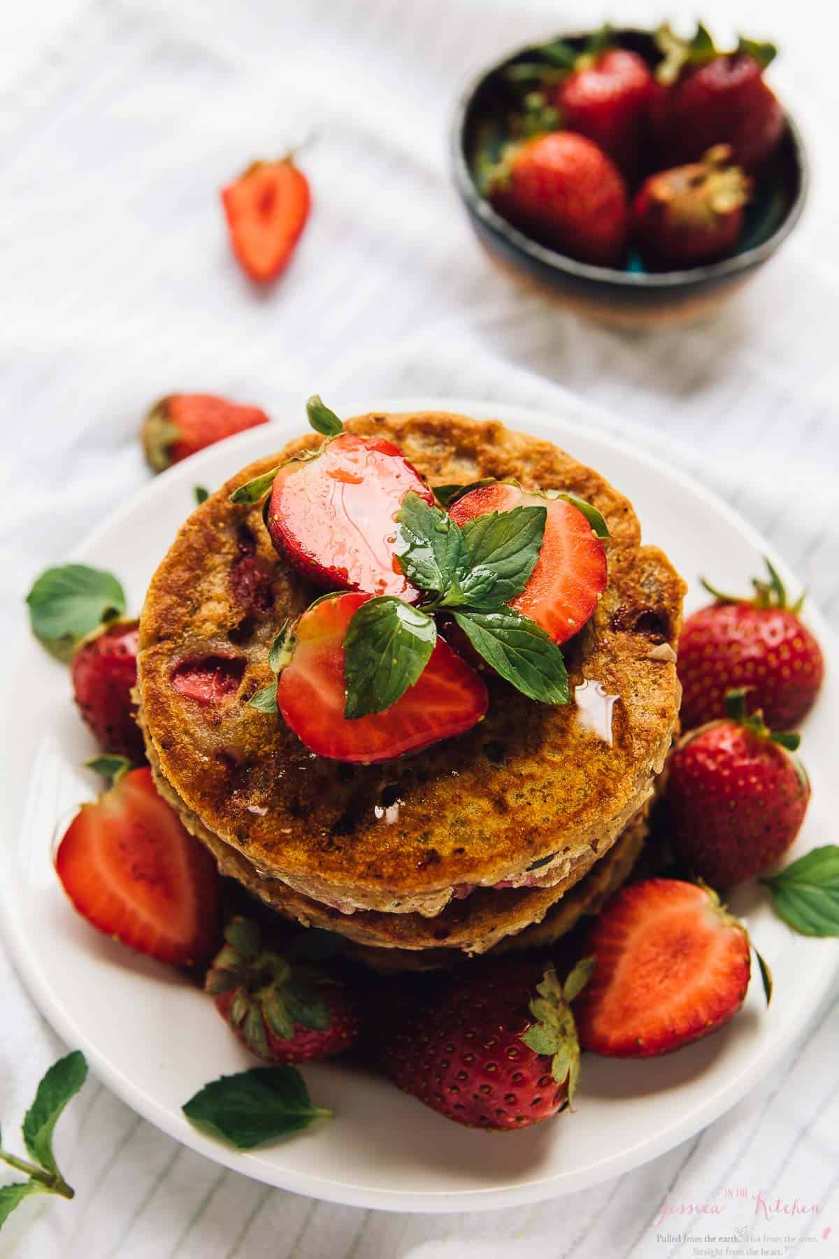 Stack of strawberry pancakes on white plate, garnished with berries and syrup