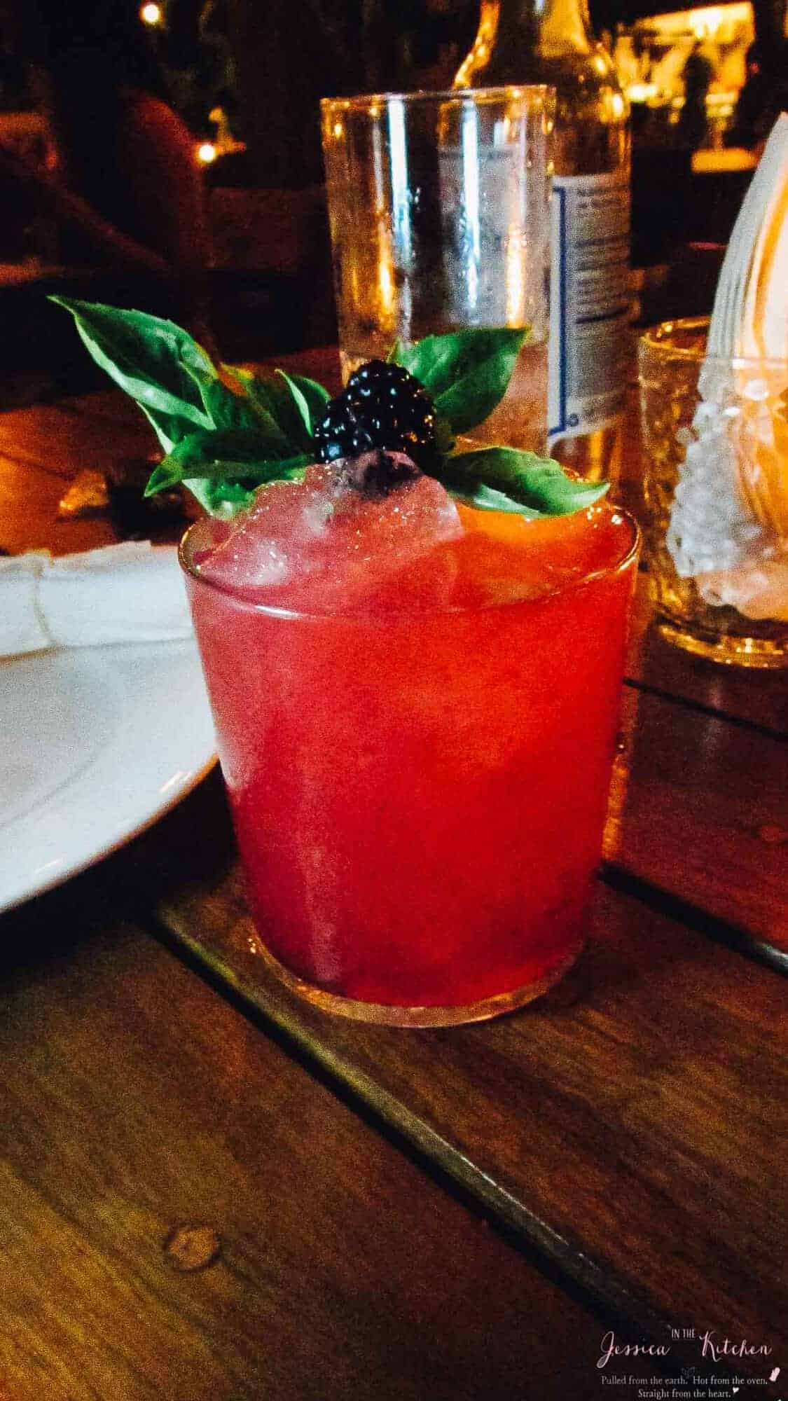 A red cocktail with blackberry garnish, on a wood table. 