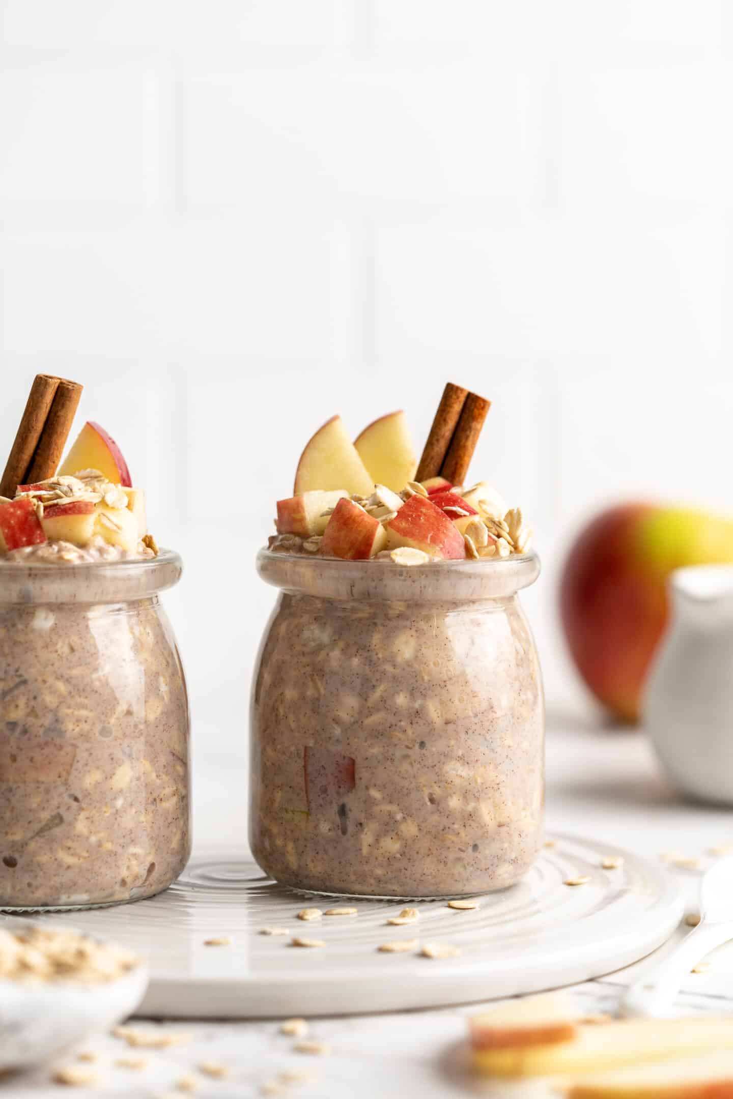 two side by side jars apple overnight oats with cinnamon sticks, oats and apples topping them