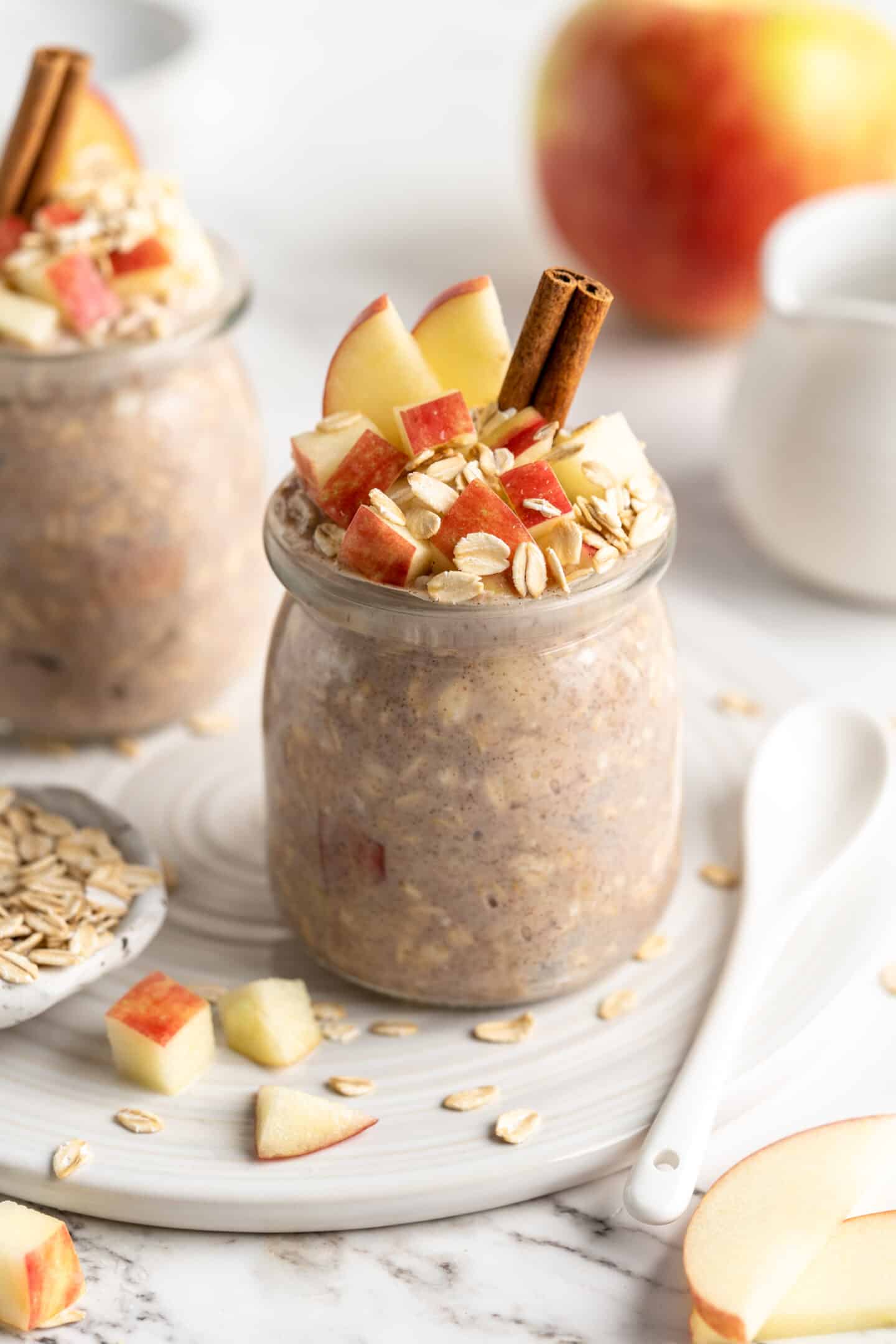 two jars apple overnight oats with cinnamon sticks, oats and apples topping them, apple in background