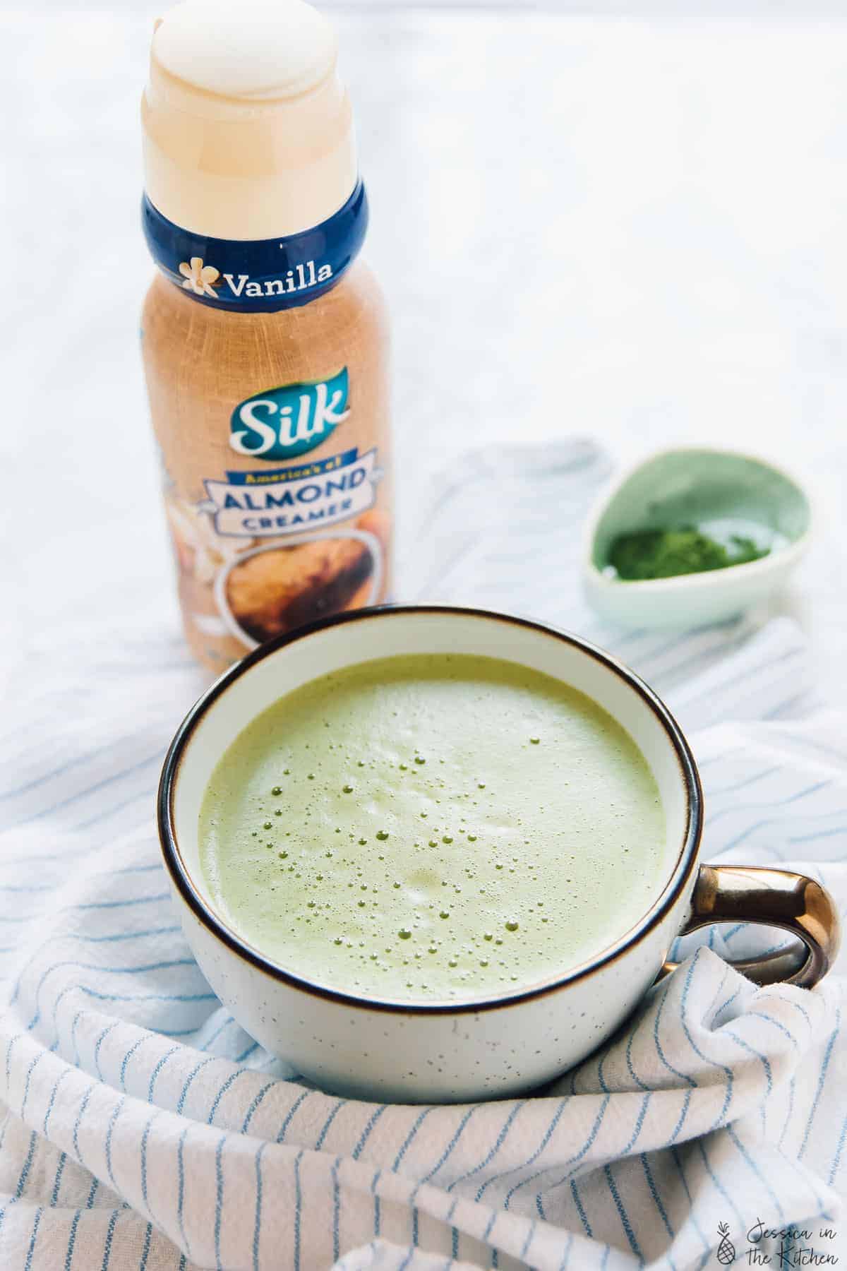 A pumpkin matcha latte in a white cup with almond creamer in the background. 