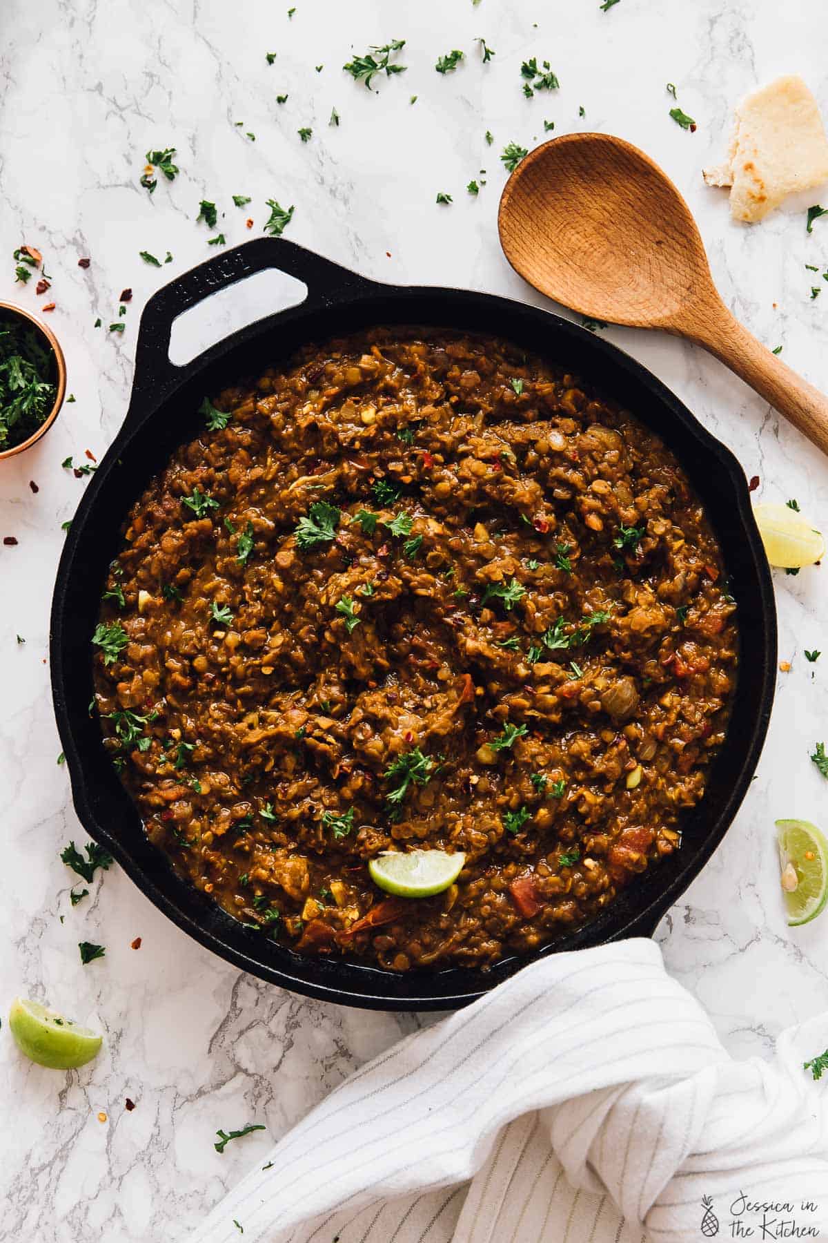 A large cast iron skillet of brown lentils garnished with parsley next to a wooden spoon