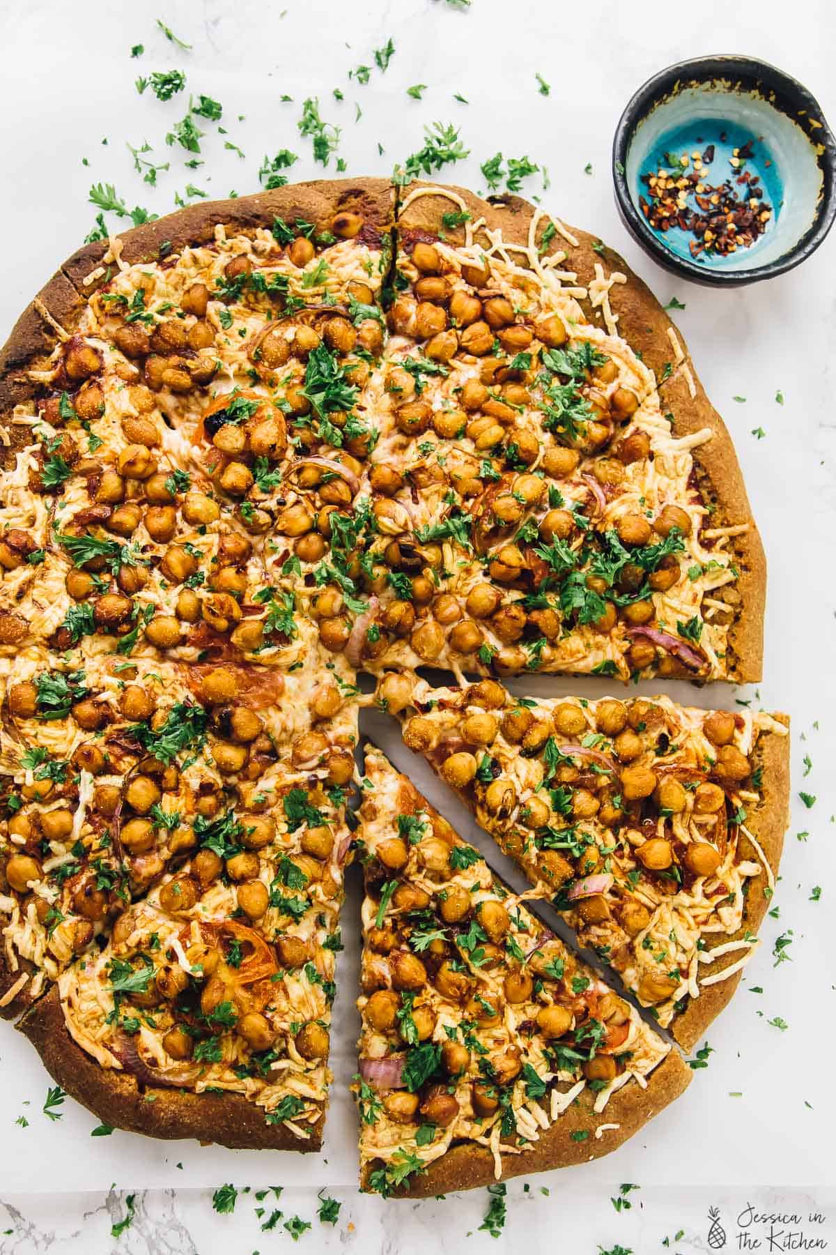 Top down shot of vegan spicy bbq chickpea pizza next to a bowl crushed red pepper flakes. 