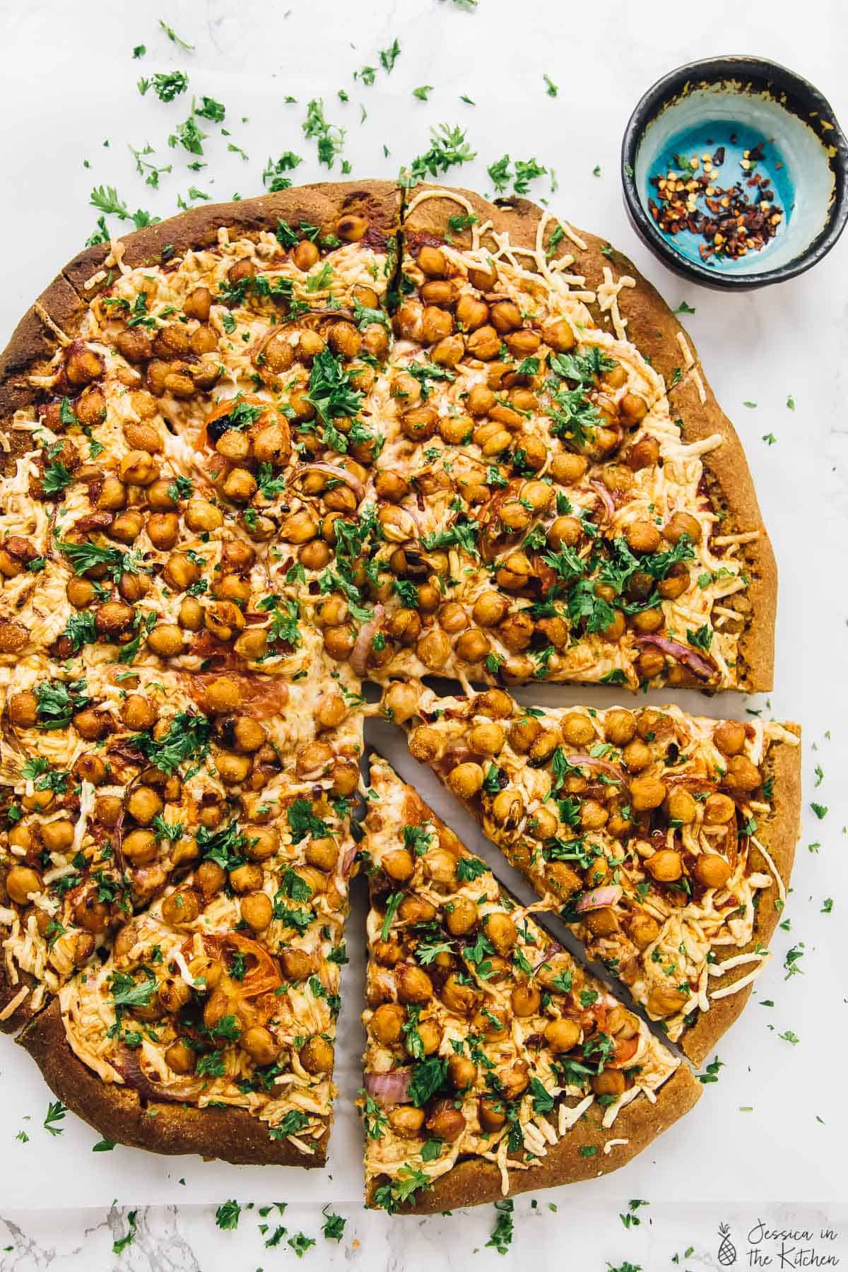 Top down view of  Vegan chickpea pizza next to a bowl of crushed red peppers. 