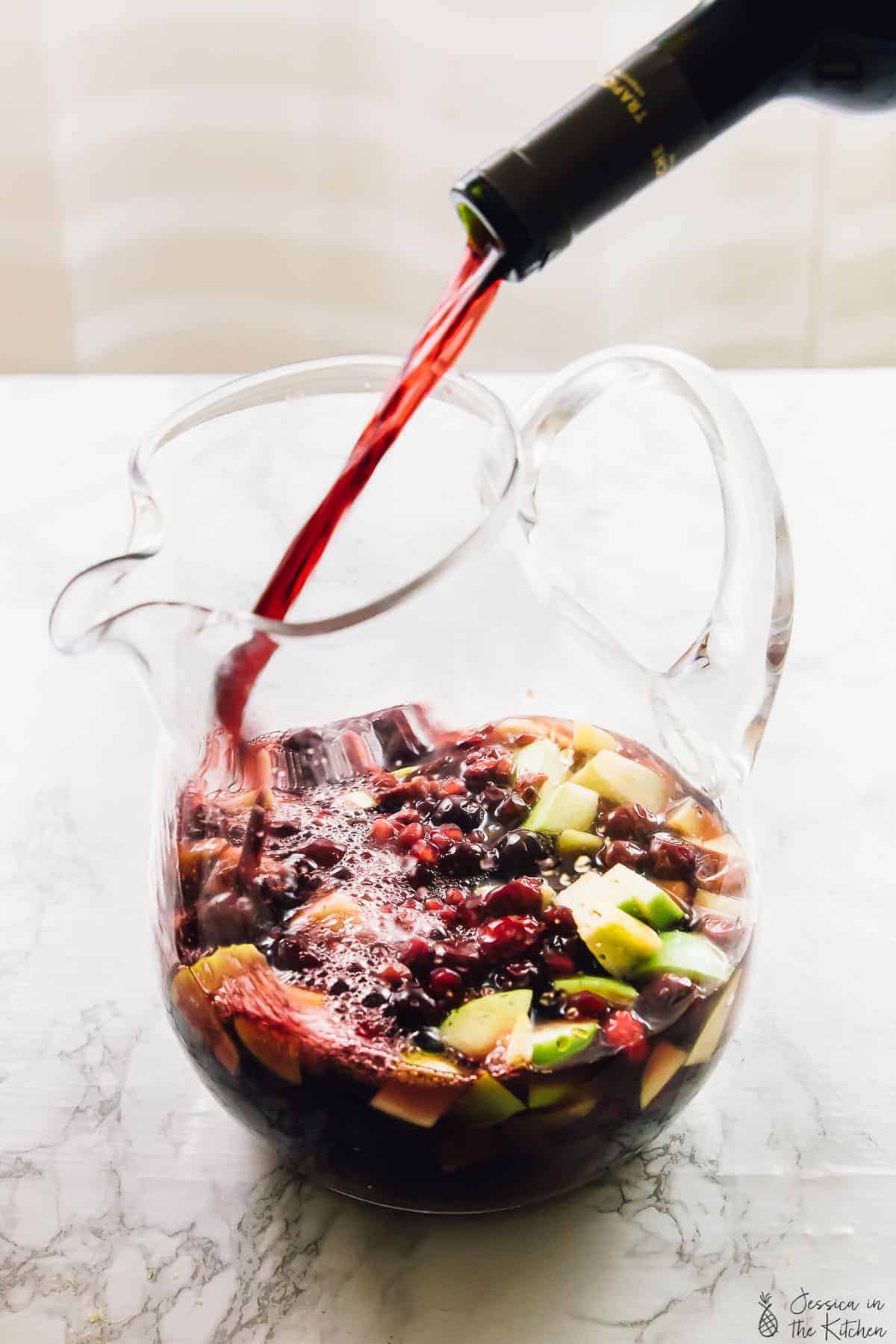 A bottle of wine being poured into a pitcher of sangria with pieces of apple, cranberries, and pomegranate arils. 