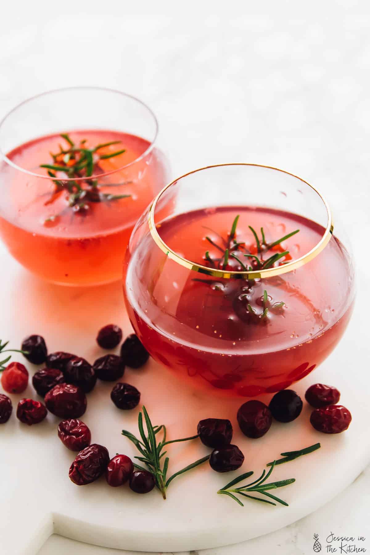 Two glasses of cranberry gin cocktail surrounded by cranberries and rosemary.
