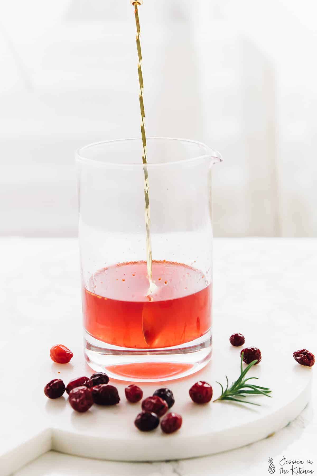 A gold spoon stirring a cranberry gin cocktail in a glass. 