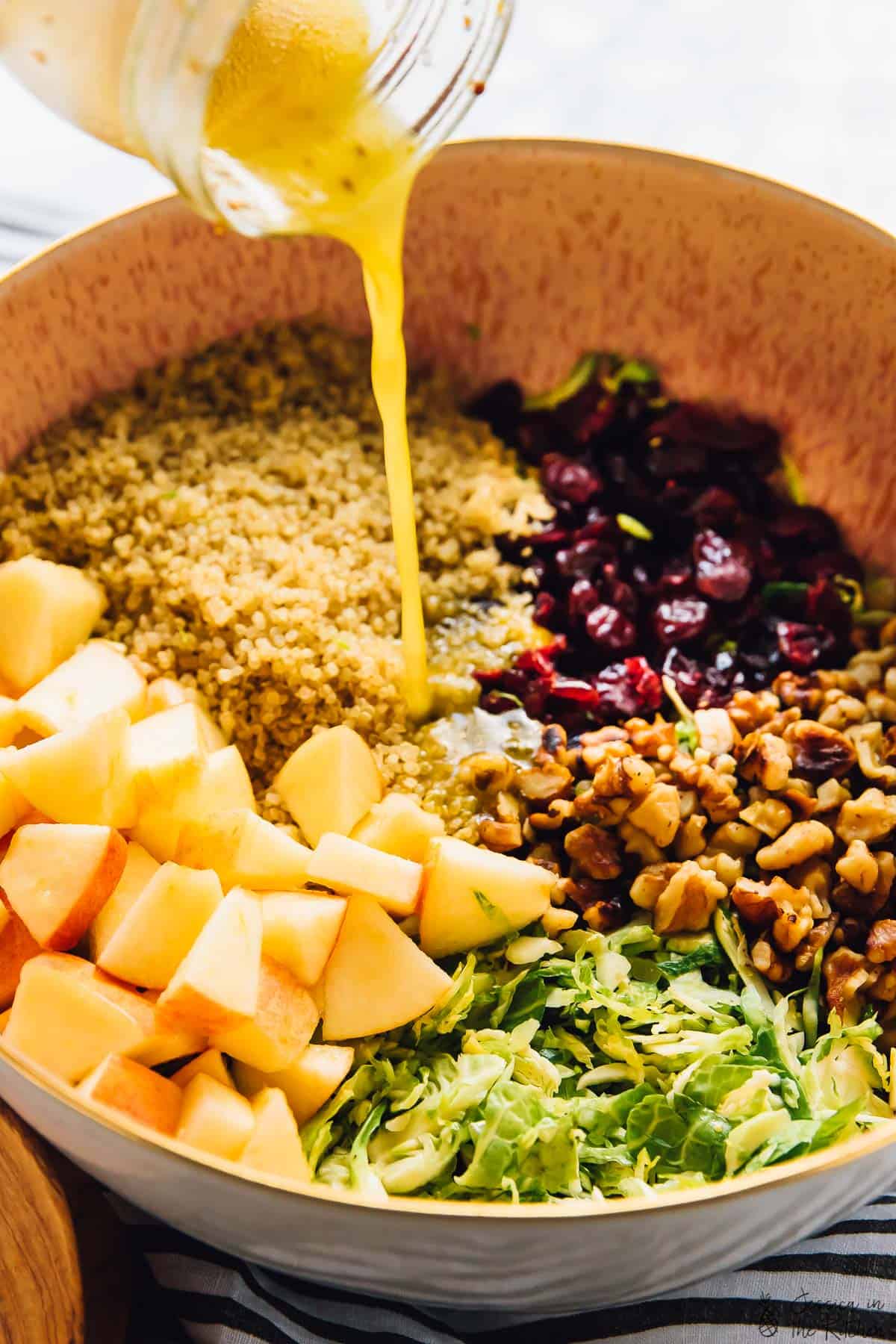 Pouring vinaigrette being poured on a shaved brussels sprouts, quinoa and apple salad in a bowl. 