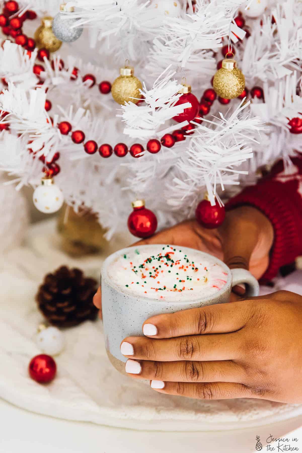 Hands holding a cup of hot chocolate under a white christmas tree. 