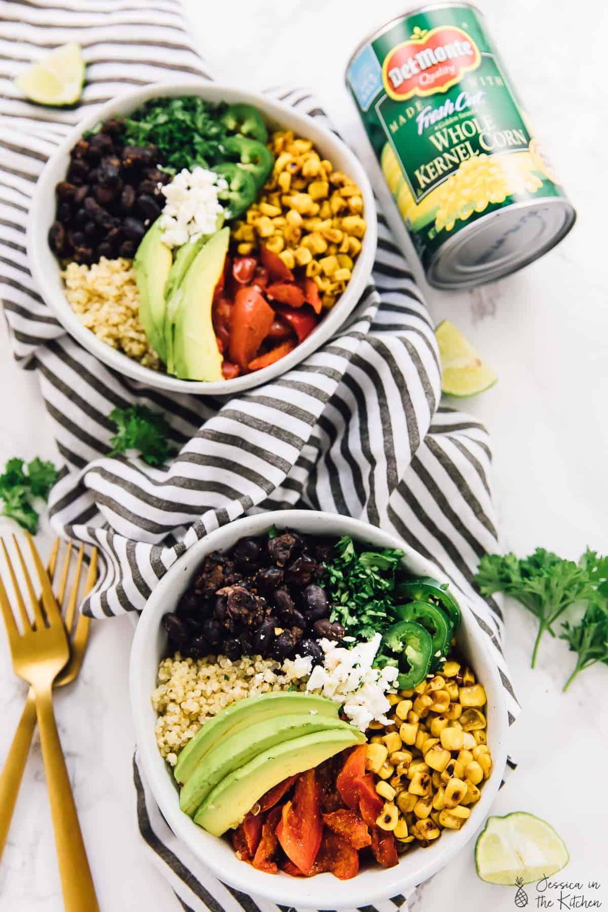Two Mexican street corn burrito bowls with two gold forks, can of corn, and garnishes on tabletop
