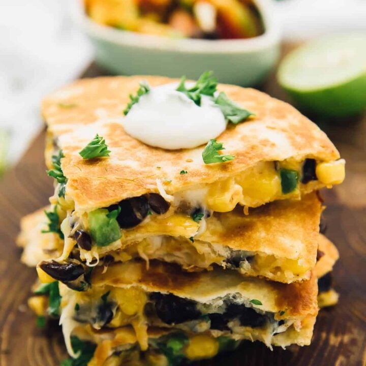 side angle view of a stack of four quesadillas on a brown cutting board with a dollop of white cream on top and parsley, with salsa and lime in the background