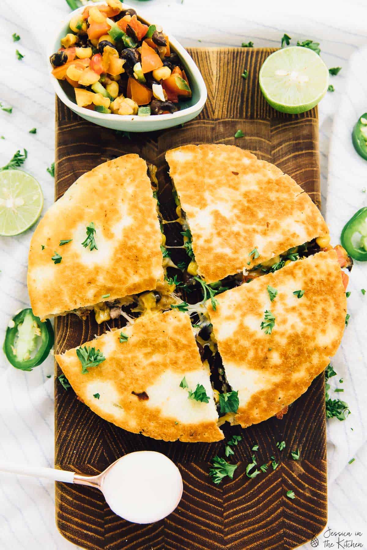 A further away shot of an overhead view of four quesadillas sliced on a brown cutting board with some cheese pull and a slight oozing of ingredients, with a sprinkle of parsley, with salsa and lime, white cream and jalapeños slices surrounding it.