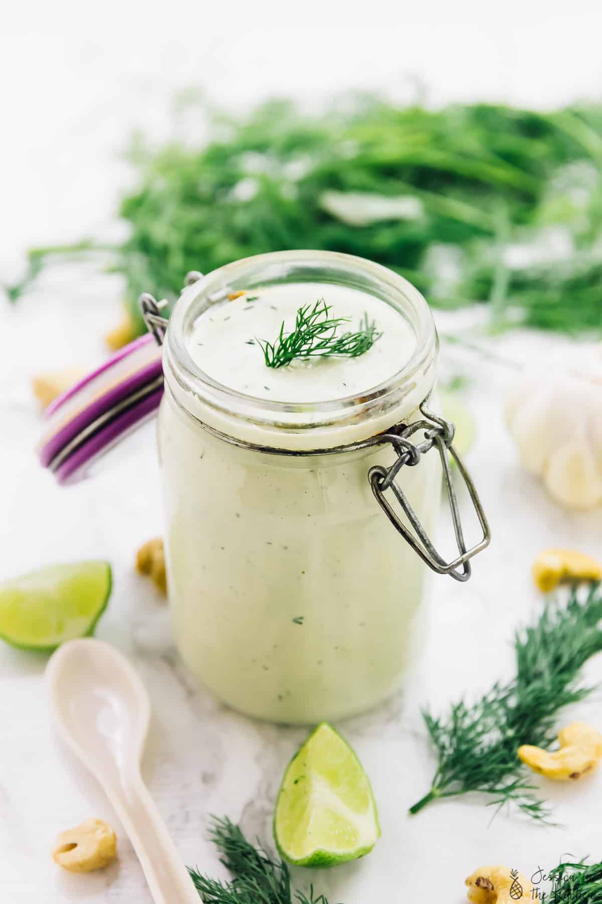 Vegan ranch dressing in a glass jar, surrounded by herbs and lime wedges. 