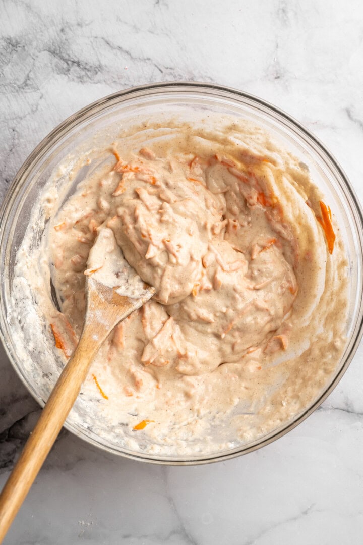 These carrot cake pancakes are fluffy, warmly spiced, and topped with an irresistible maple cream cheese drizzle. A cozy vegan breakfast! Carrot cake pancake batter in glass bowl with wooden spoon.