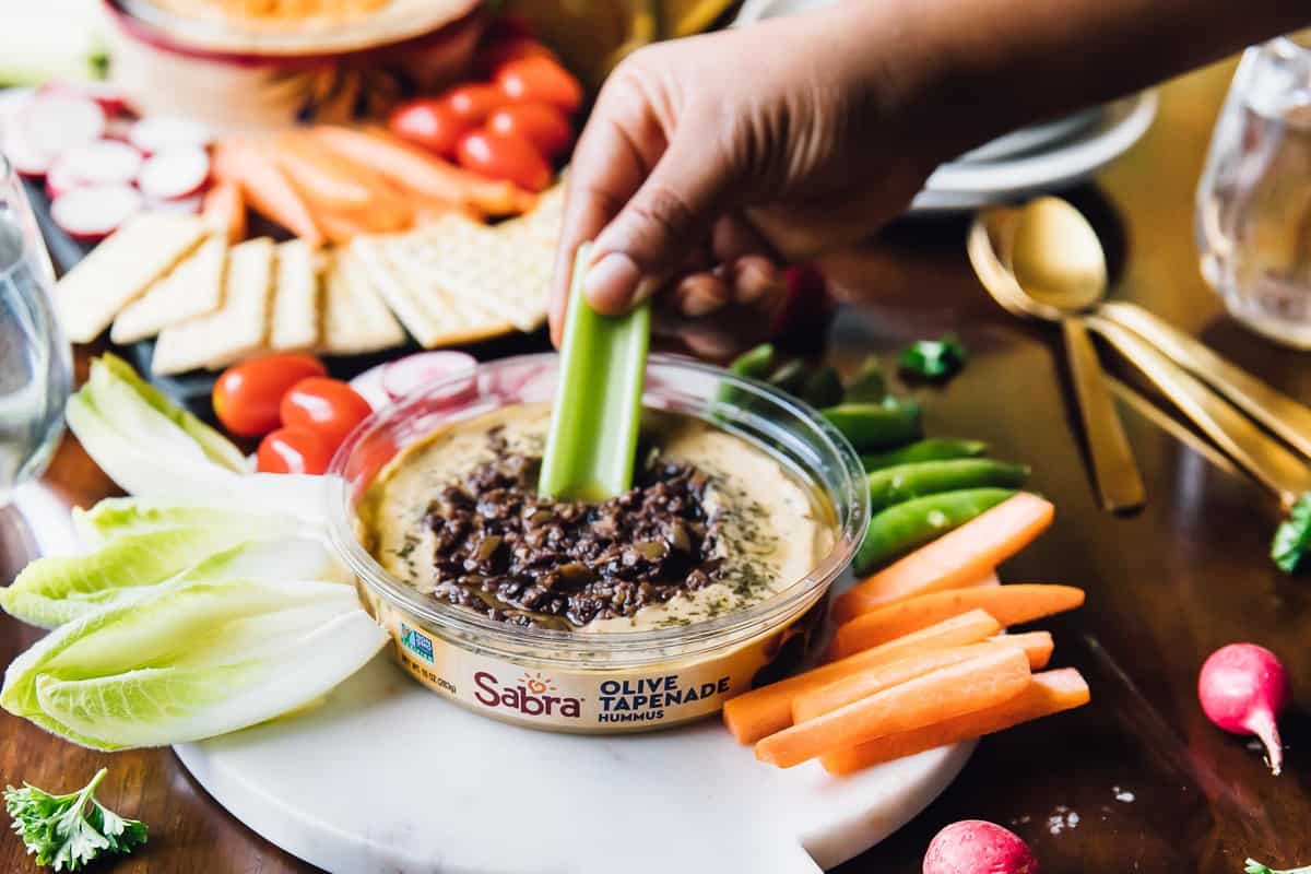 Hand dipping a celery stick into some tapenade, surrounded by veggies. 
