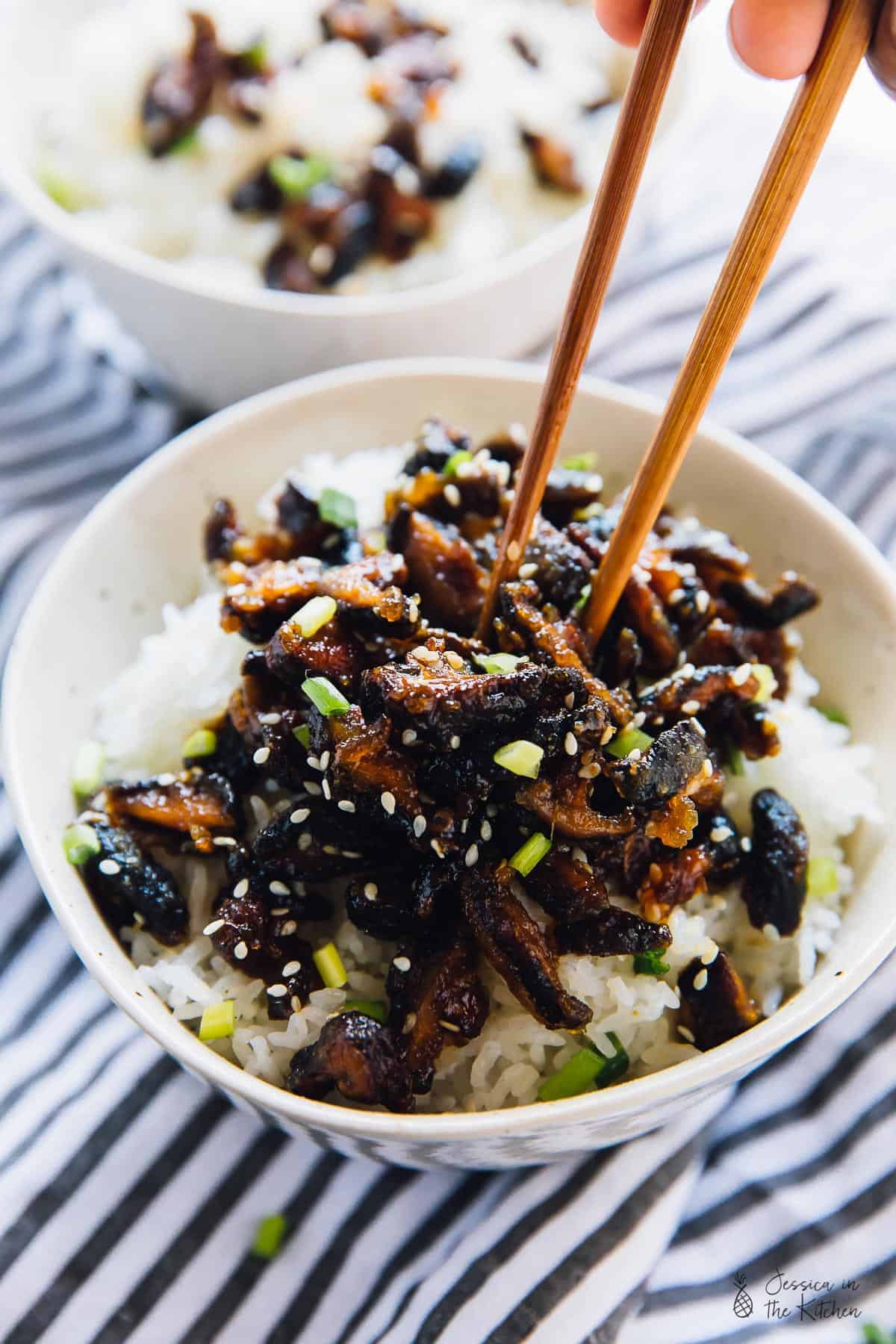 Chopsticks digging into a bowl of sticky sesame shiitake mushrooms for a roundup on Vegan Takeout Recipes.