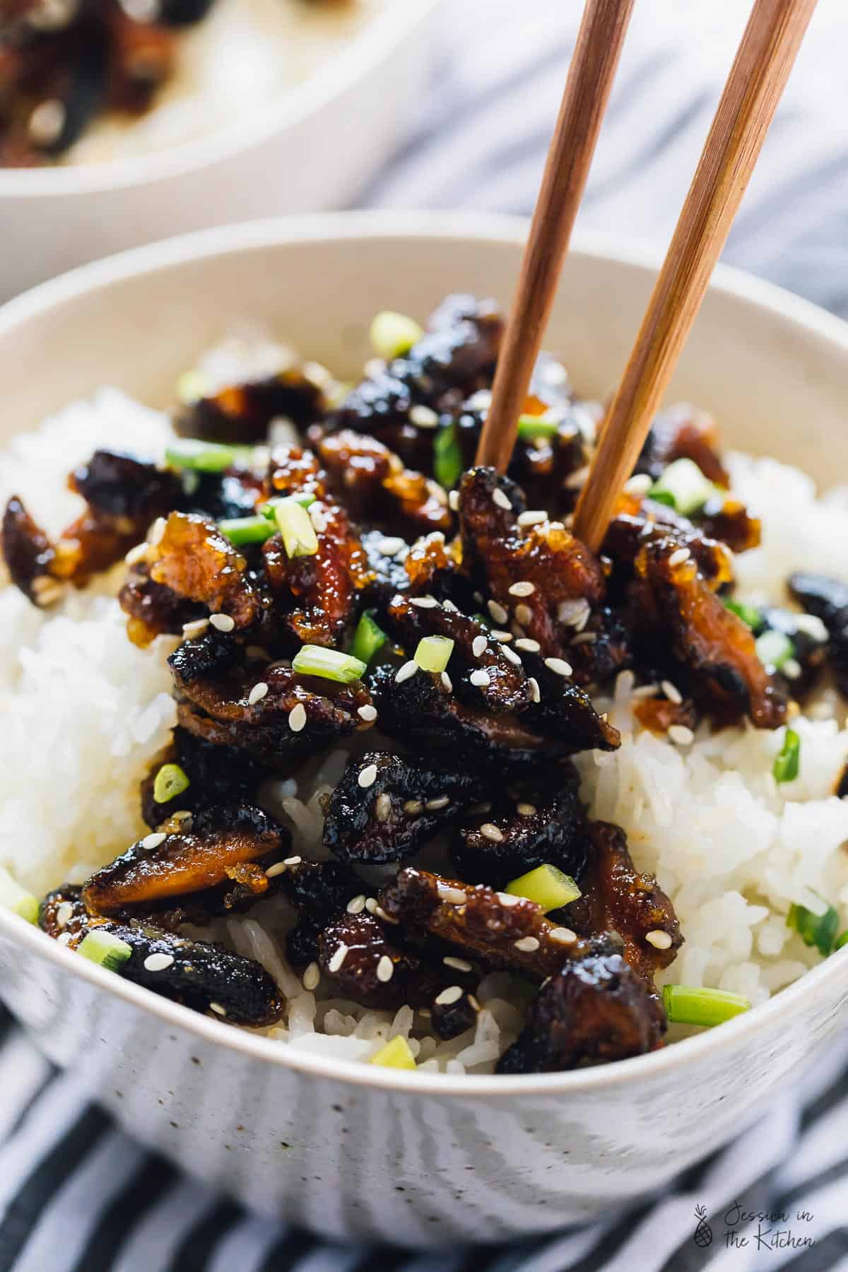 Sticky sesame shiitake mushrooms on rice, in a white bowl with chopsticks.