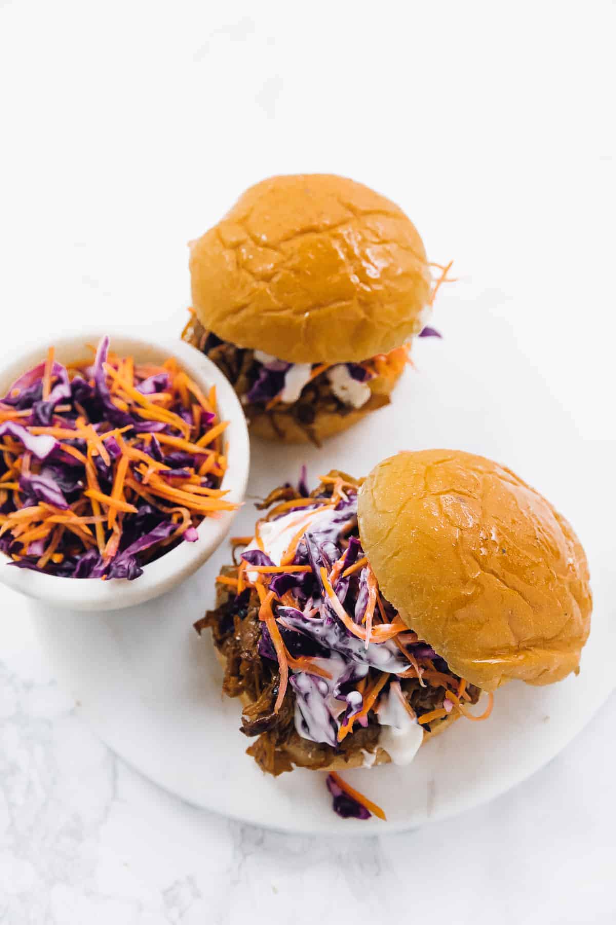 Top down view of two vegan pulled mushroom sandwiches on a white plate with a bowl of slaw on the side. 