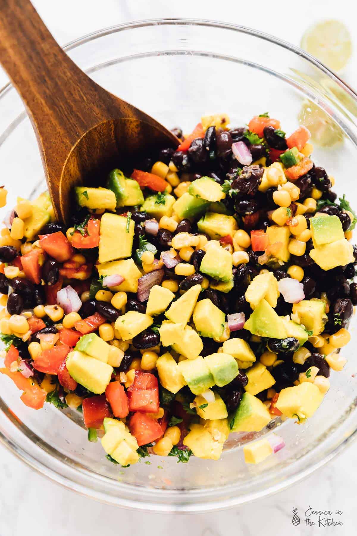 A wooden spoon stirring black bean avocado and corn salad in a glass bowl. 