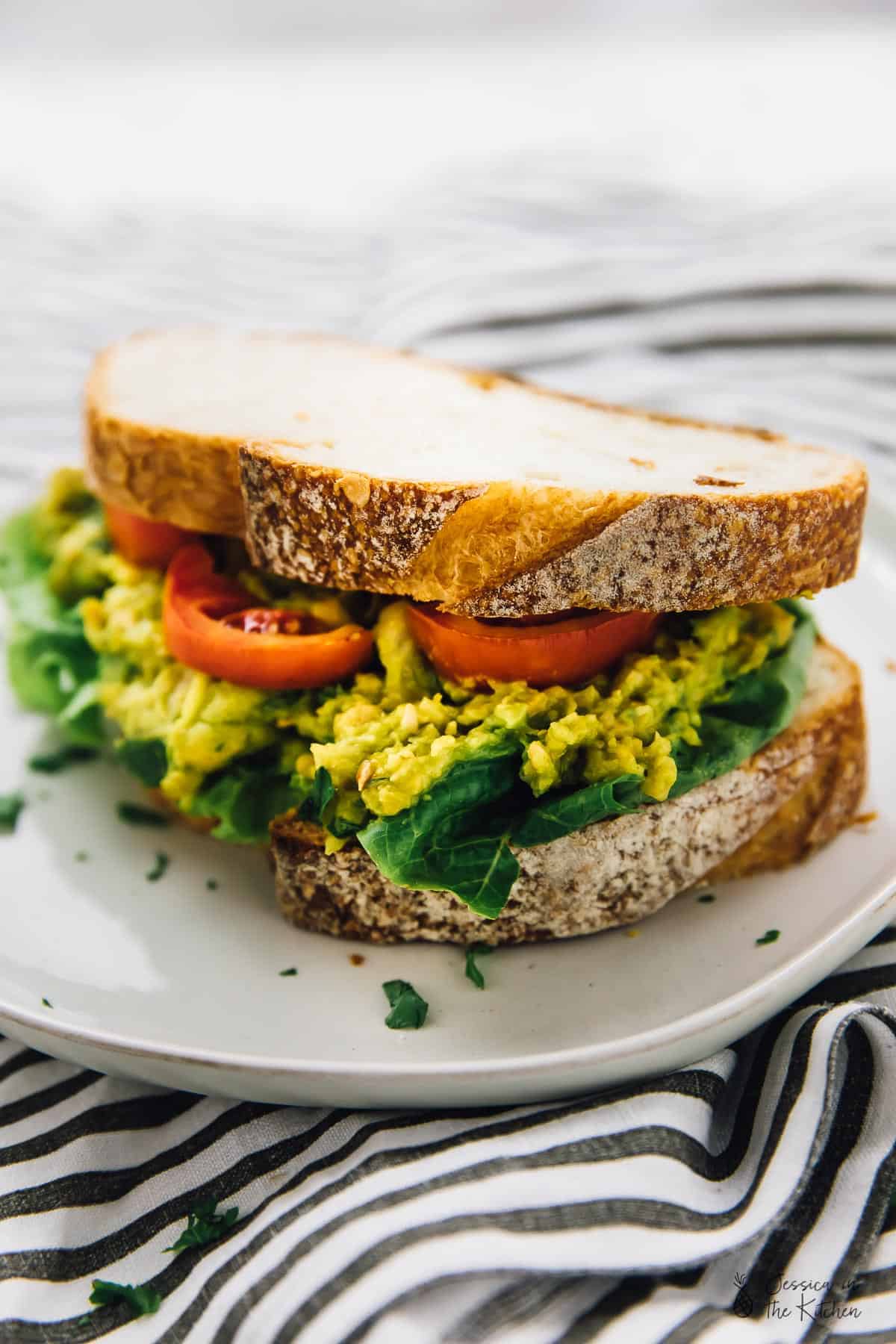 A smashed chickpea avocado sandwich with sliced tomato, on a plate. 