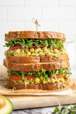 Two smashed chickpea avocado salad sandwiches stacked on cutting board