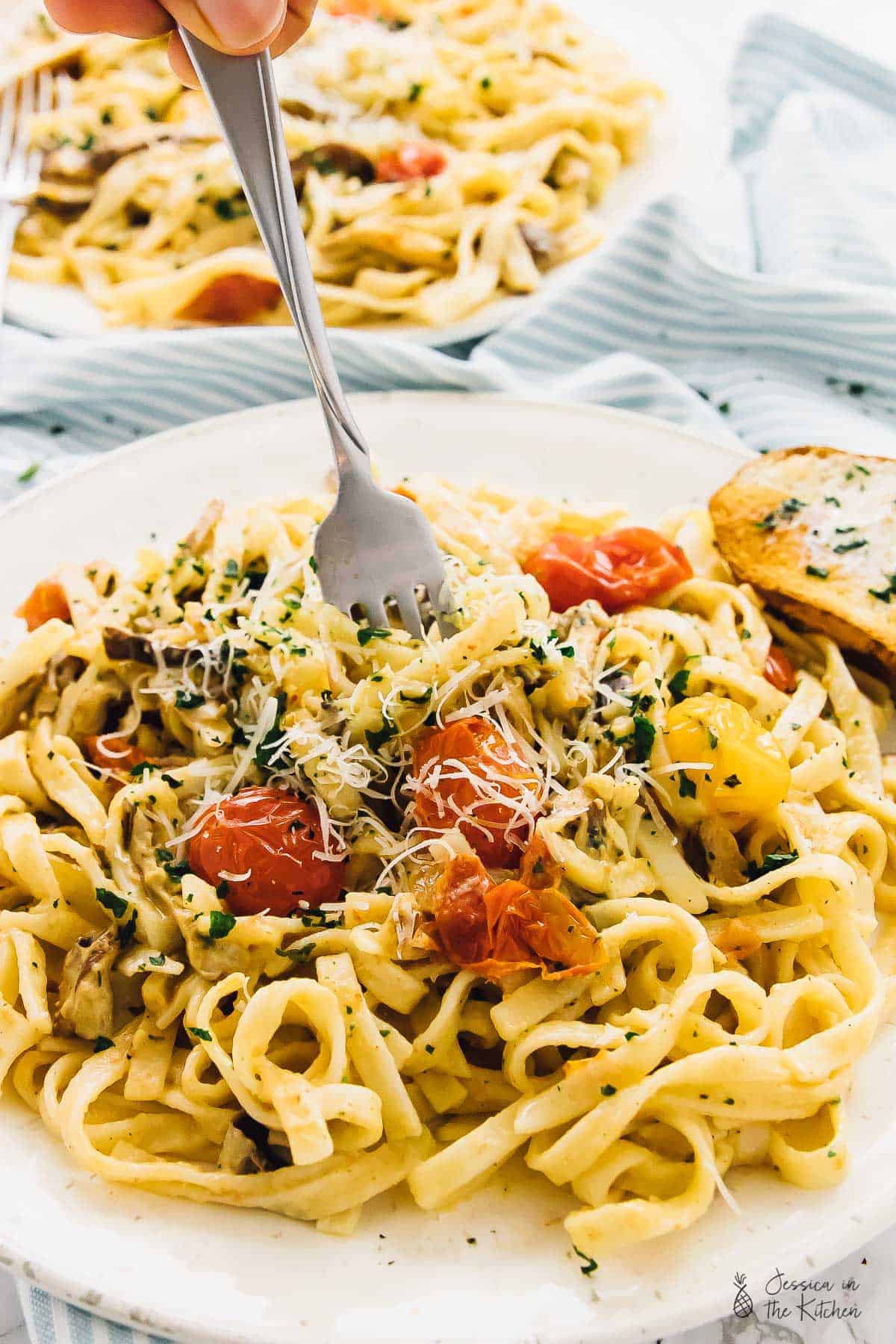 A fork in a plate of fettuccine pasta with burst tomatoes, fresh herbs, and Parmesan cheese