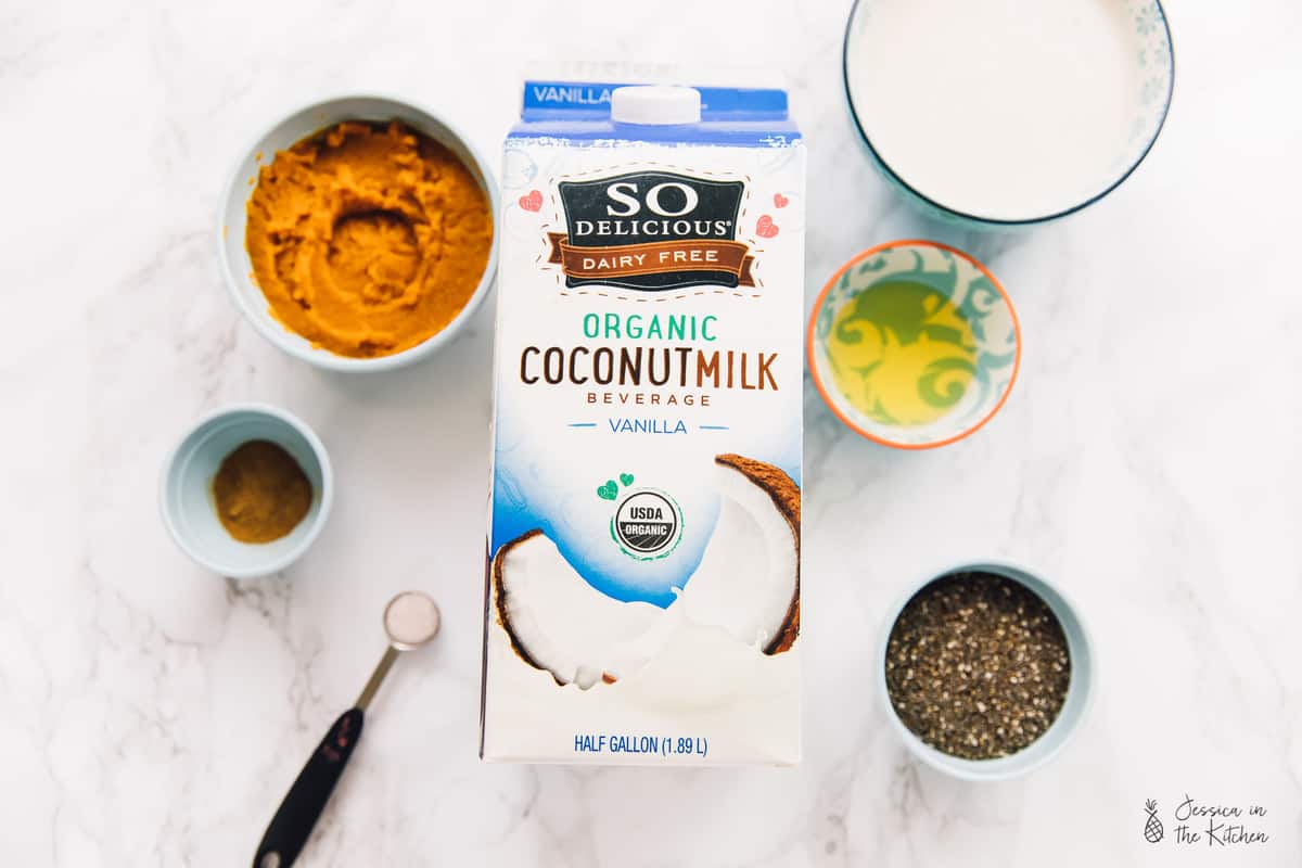 Overhead view of the ingredients needed for pumpkin chia pudding: a carton and a bowl of of coconut milk beverage, a bowl of pumpkin puree, a bowl of agave, a bowl of chia seeds, a bowl of pumpkin pie spices, and a measuring spoon of salt