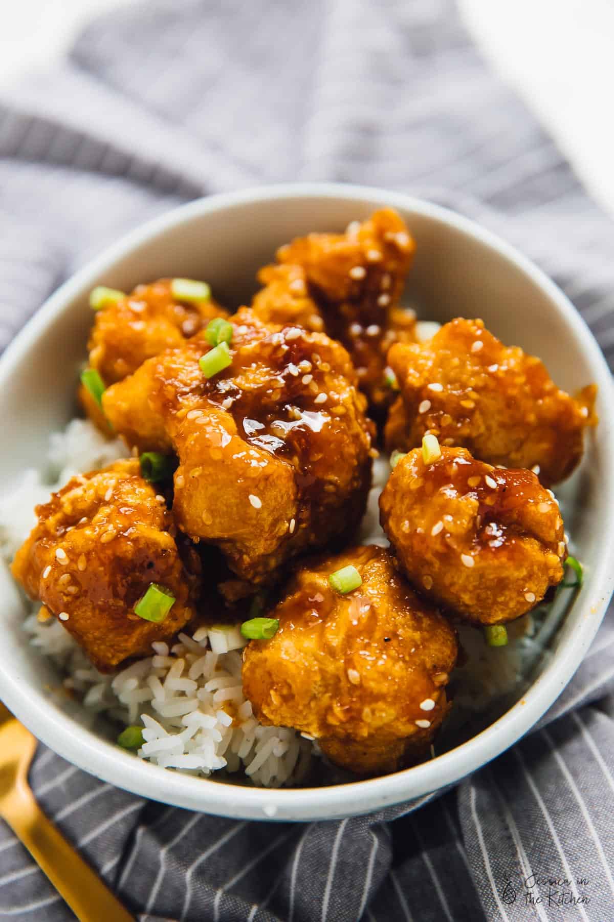 A bowl of sweet and sticky orange cauliflower bites on a bed of rice, in a grey bowl. 