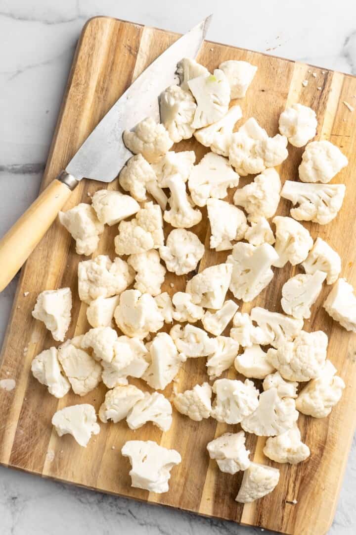 Overhead view of cauliflower cut into florets on wooden board with knife