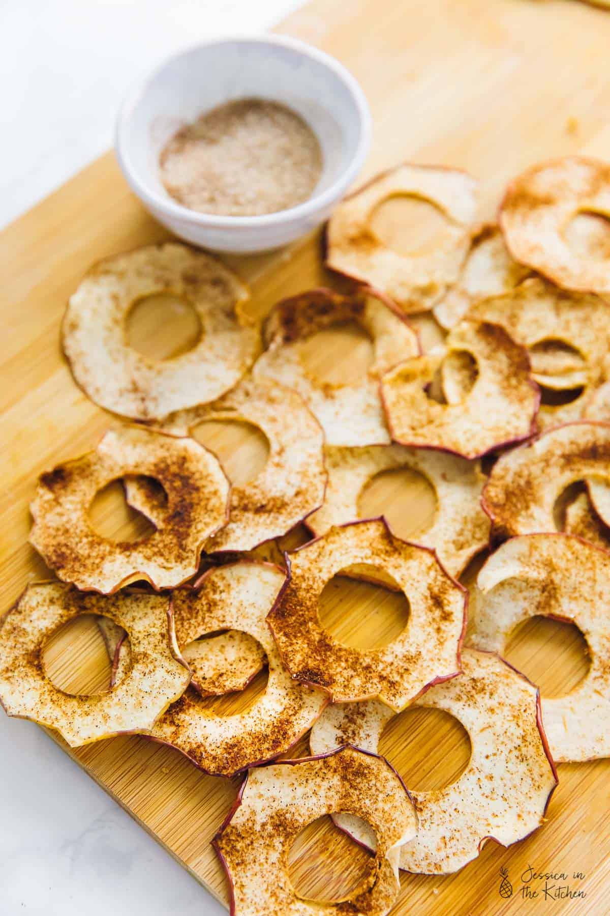 Overhead view of a bunch of apple chips on a cutting board, covered in cinnamon, next to a bowl of cinnamon sugar. 