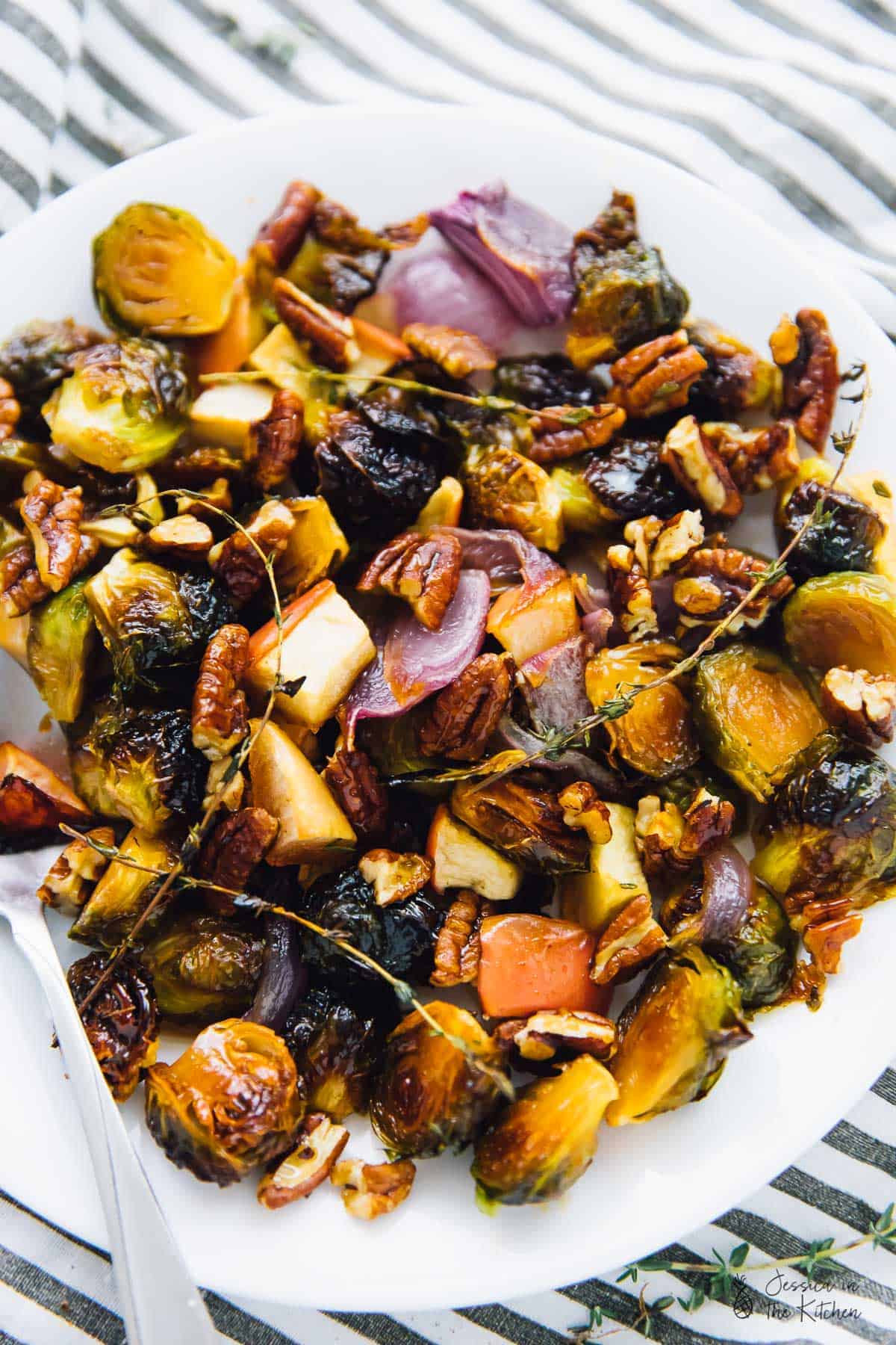 A dish full of roasted Brussels sprouts, apples, onions, and thyme, topped with pecans, with a serving spoon. 