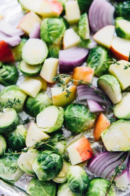 Close up of raw Brussels sprouts, apples, onions, and thyme on a baking sheet.