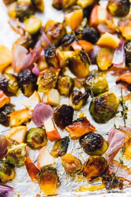 Close up of roasted Brussels sprouts, apples, onions, and thyme on an aluminum foil-lined baking sheet. 