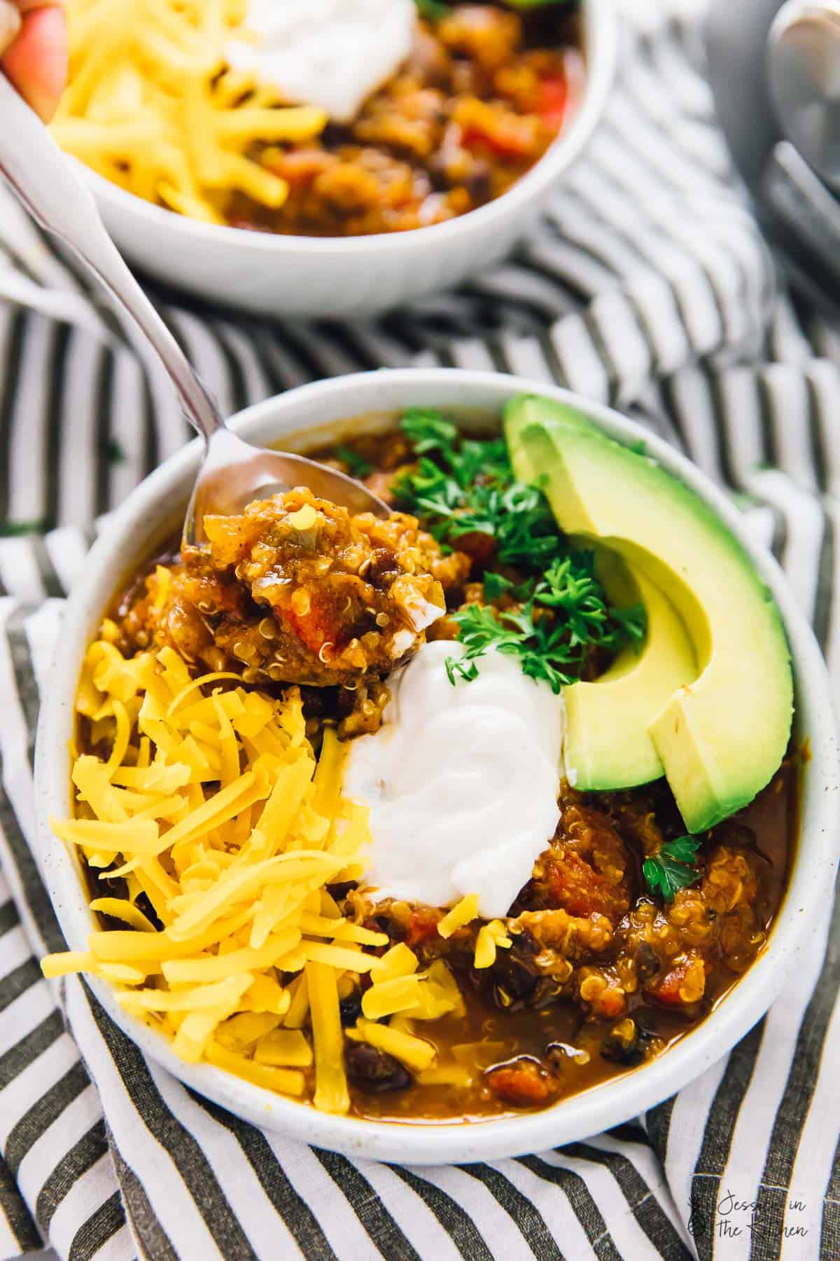 Slow cooker pumpkin quinoa chili in a white bowl, topped with sliced avocado, cheese and cream. 