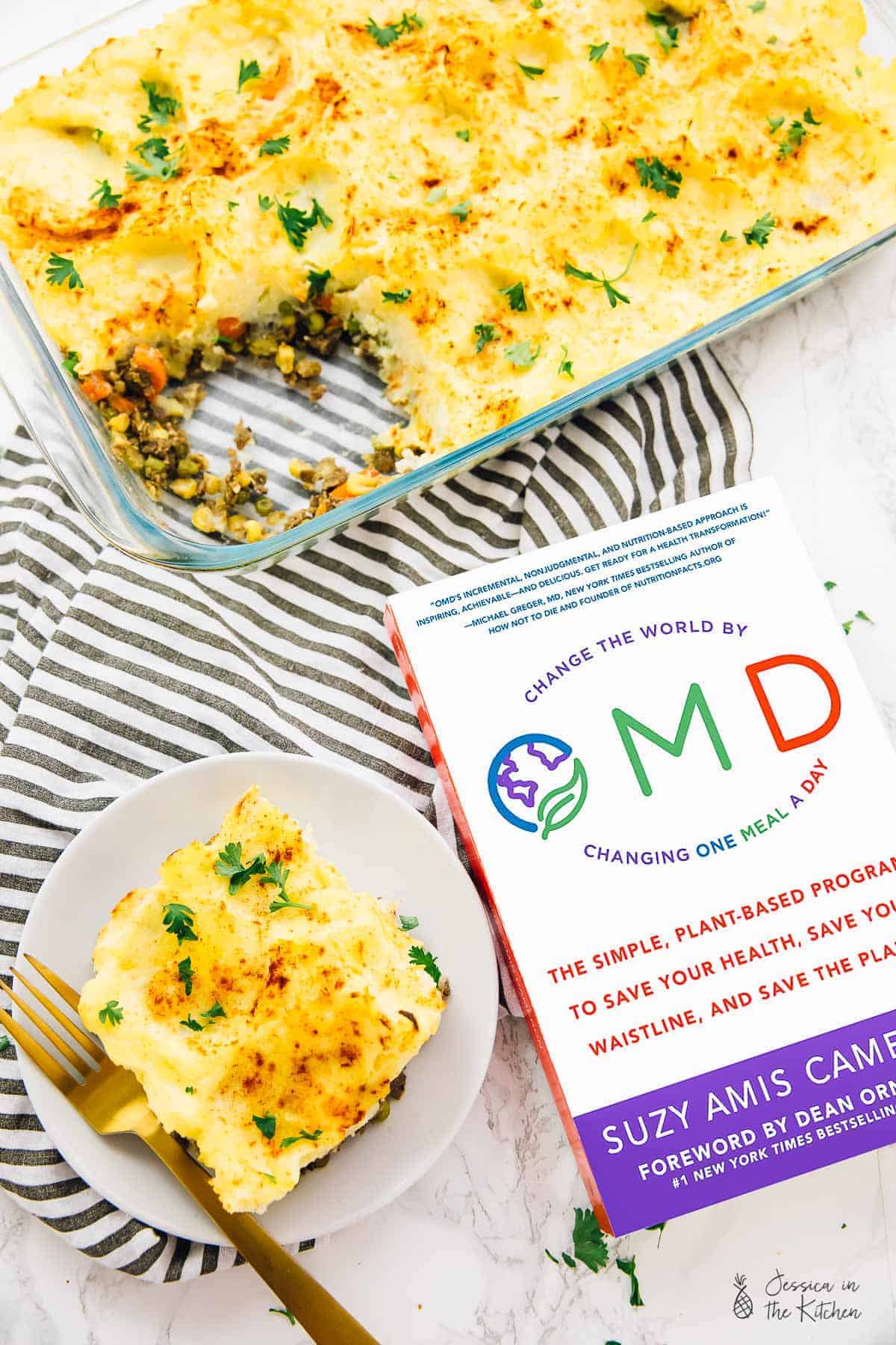 Vegan shepherd's pie in a baking dish, next to a portion of pie on a plate and a cook book. 