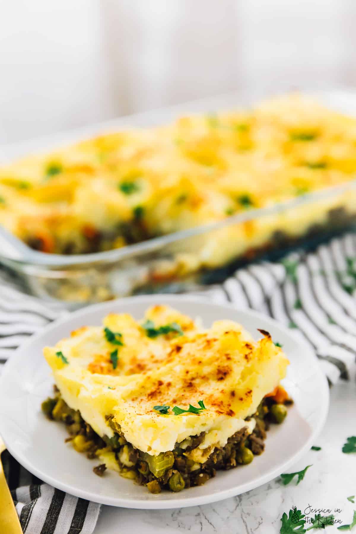 A portion of vegan shepherd's pie on a plate, with a baking dish shepherd's pie in the background.