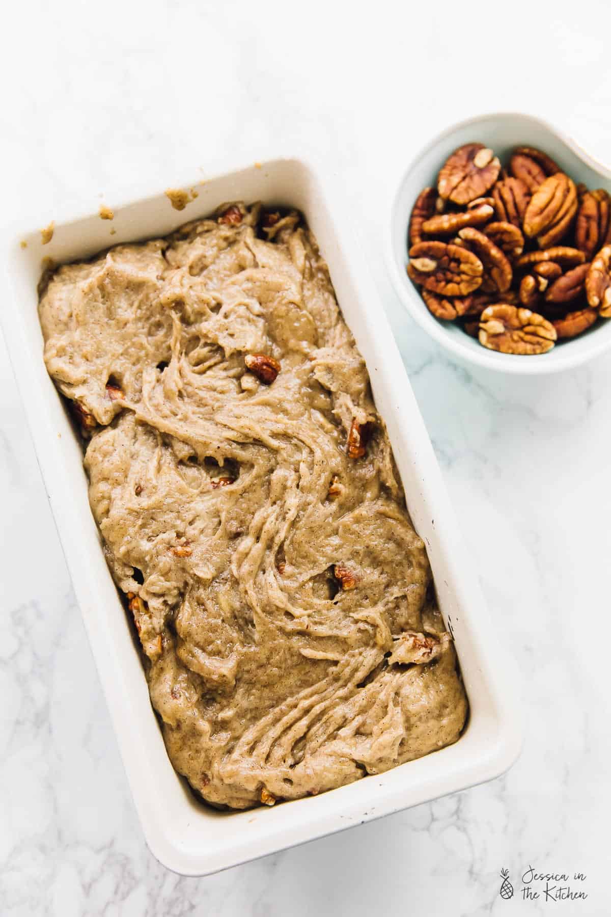 Banana bread batter in a baking dish, next to a bowl of pecans. 