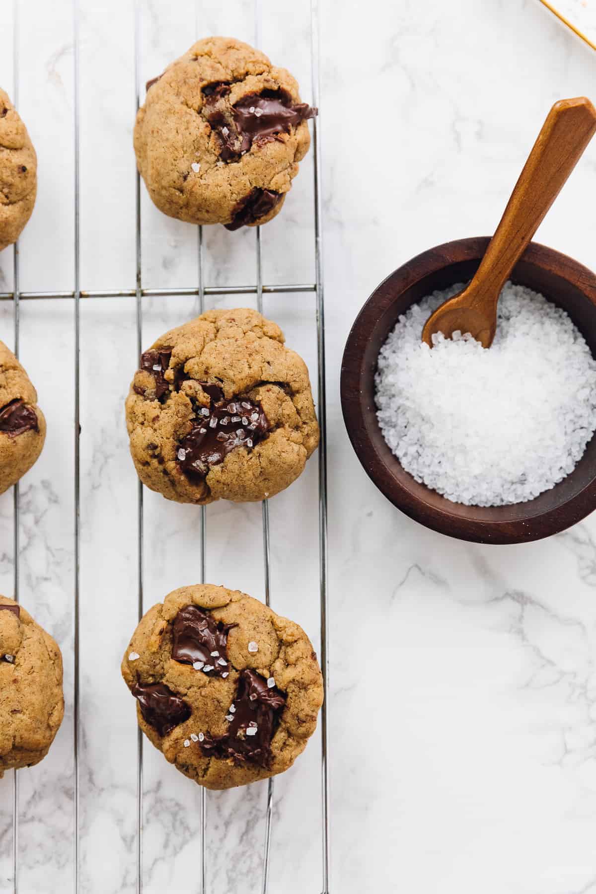 Salted chocolate chip tahini cookies on a wire rack next to bowl of flaky sea salt