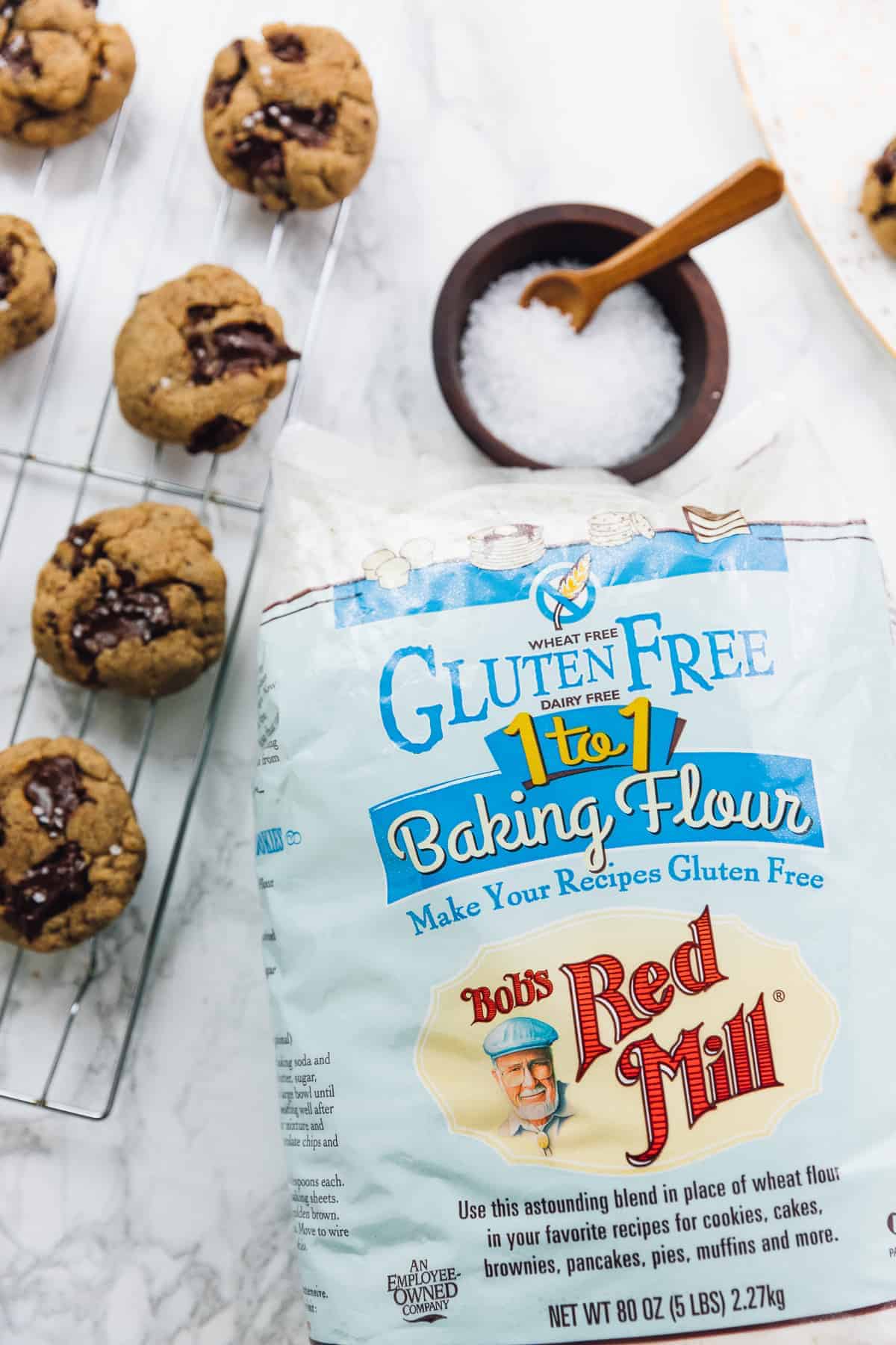 Salted chocolate chip tahini cookies on a wire rack, next to a pack of baking flour.