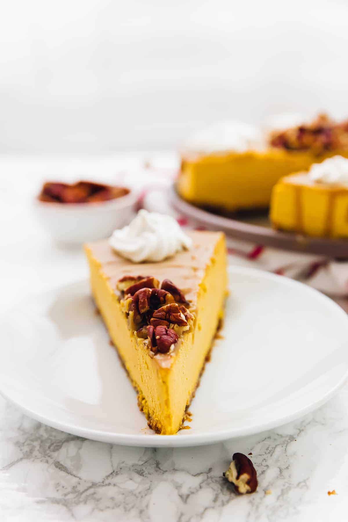 A slice of pumpkin cheesecake on a plate, topped with nuts and whipped cream, with a plate of cake in the background