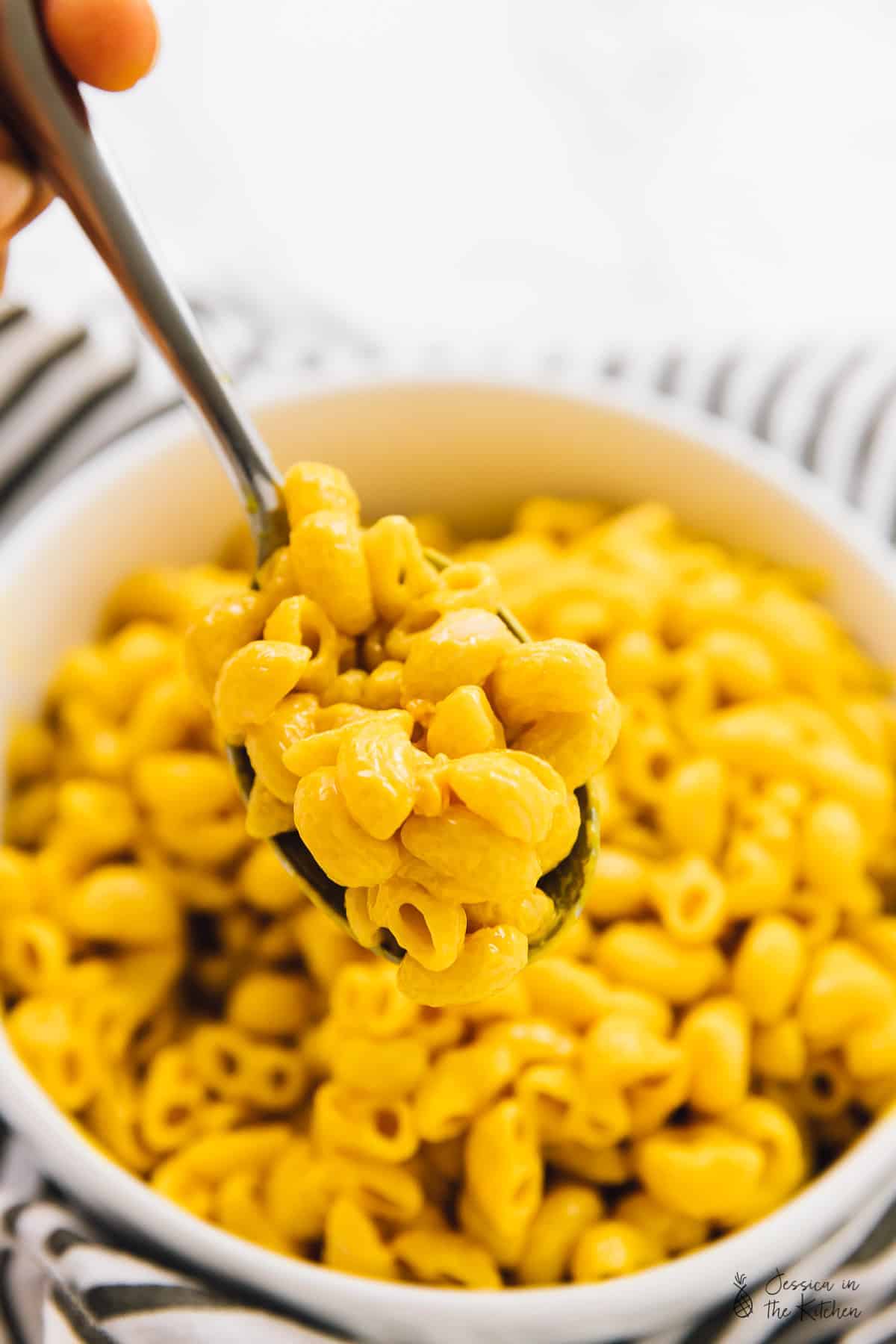 A hand holding a spoonful of mac and cheese, with a bowl of mac and cheese in the background. 