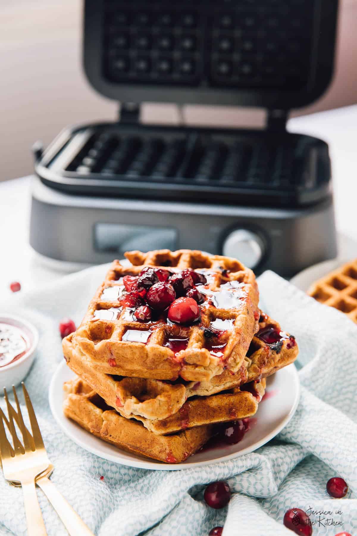 A stack of orange cranberry waffles in front of a waffle iron. 