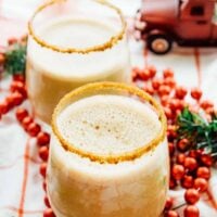 Two sugar rimmed glasses of vegan eggnog on a table with holiday decorations.