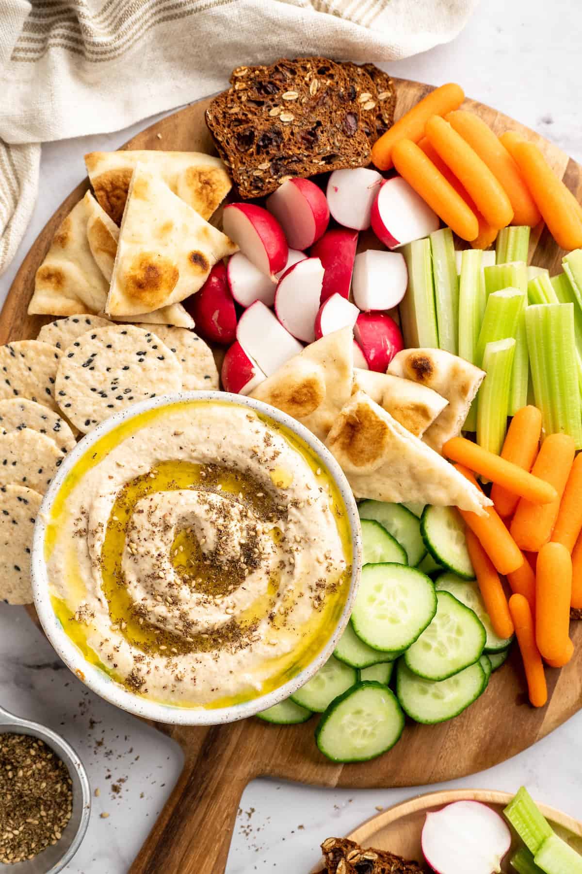 This easy Roasted Cauliflower Hummus gives the classic dip a low-carb makeover! I add roasted garlic too, which makes it extra delicious. Overhead view of roasted cauliflower hummus in bowl on board with bread, crackers, and veggies