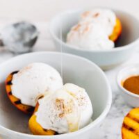 Grilled peaches and coconut ice cream in a bowl with maple syrup drizzled on top.