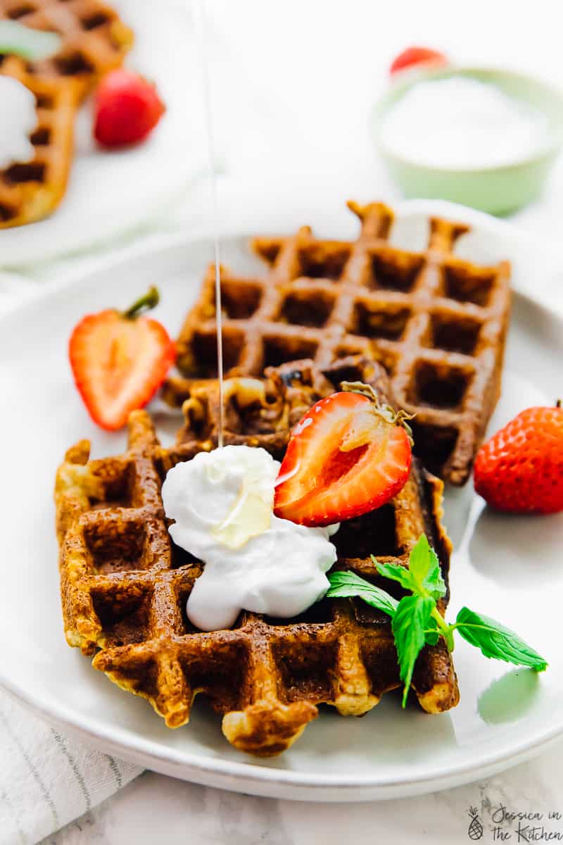 Two low carb waffles with coconut whipped cream a strawberry and a mint leaf on a plate surrounded by strawberries.