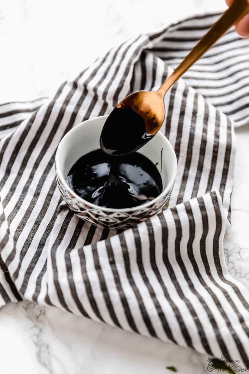 Small bowl of balsamic glaze on striped cloth napkin with spoon dripping glaze into bowl