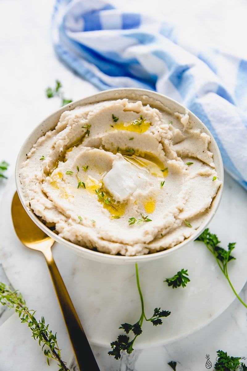 A bowl of cauliflower mash next to a blue cloth, with a gold spoon and sprigs of fresh herbs